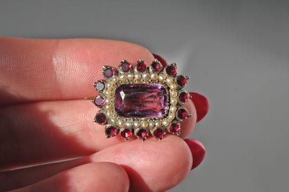 An antique Georgian era brooch with a rectangular purple amethyst, a halo of seed pearls and red flat or table cut garnets, held in someone's hand 