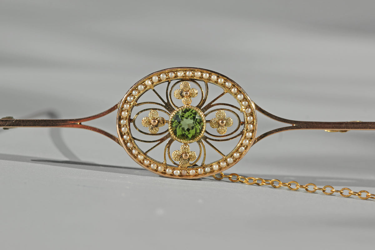 A close-up of an ornate 9ct gold edwardian jewelry brooch with a circular filigree design, a central green peridot gemstone, small pearls around the oval, and a gold safety chain extending to the right, photographed on a light gray background.