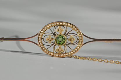 A close-up of an ornate 9ct gold edwardian jewelry brooch with a circular filigree design, a central green peridot gemstone, small pearls around the oval, and a gold safety chain extending to the right, photographed on a light gray background.