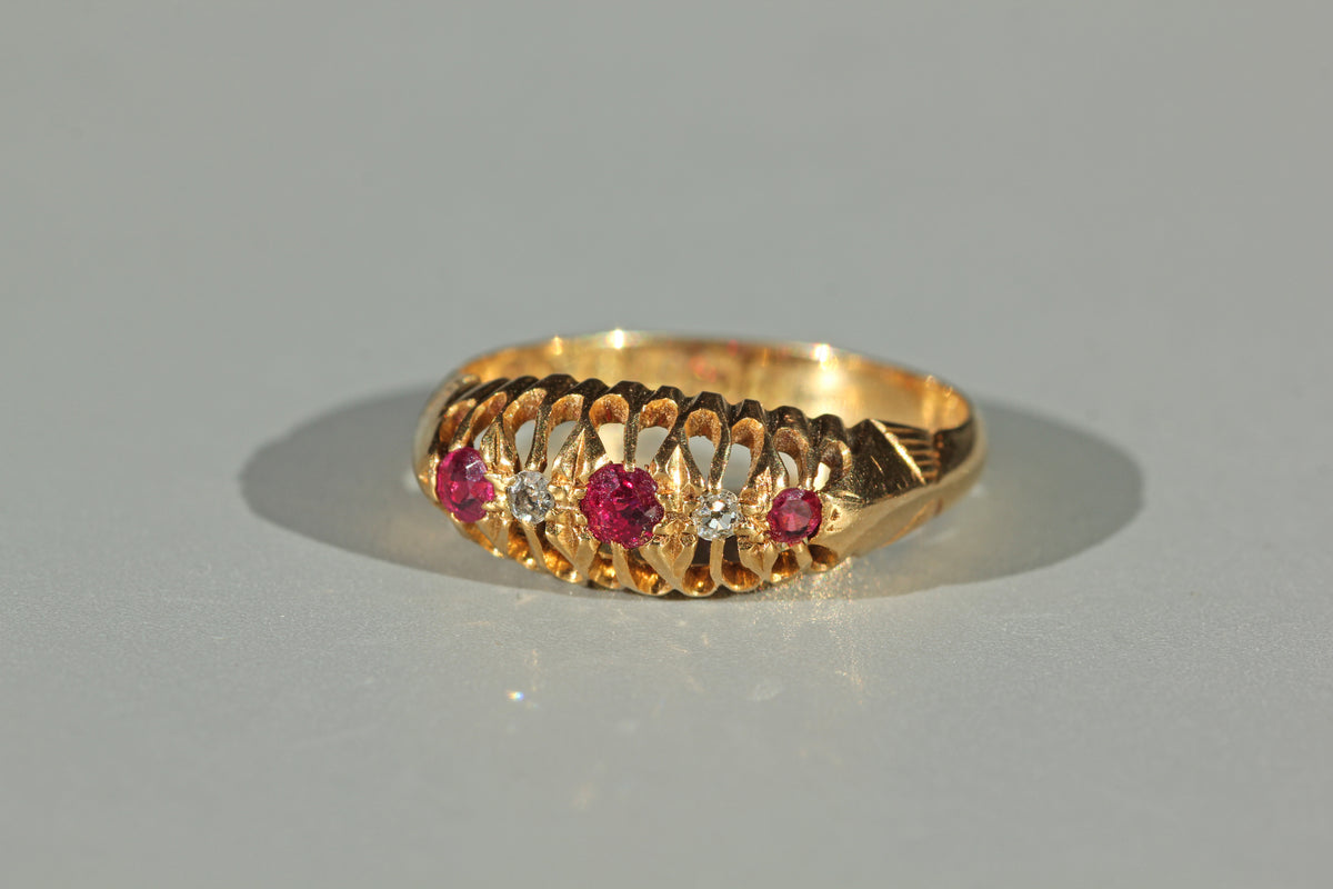 A yellow 18ct gold ring featuring a decorative openwork gallery and a row of pink ruby gemstones with small clear diamond stones, photographed on a neutral gray background.