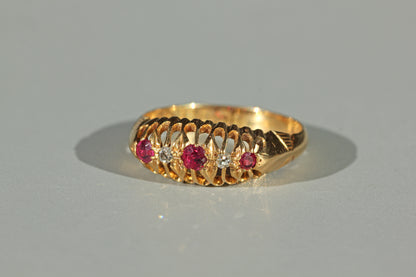A yellow 18ct gold ring featuring a decorative openwork gallery and a row of pink ruby gemstones with small clear diamond stones, photographed on a neutral gray background.