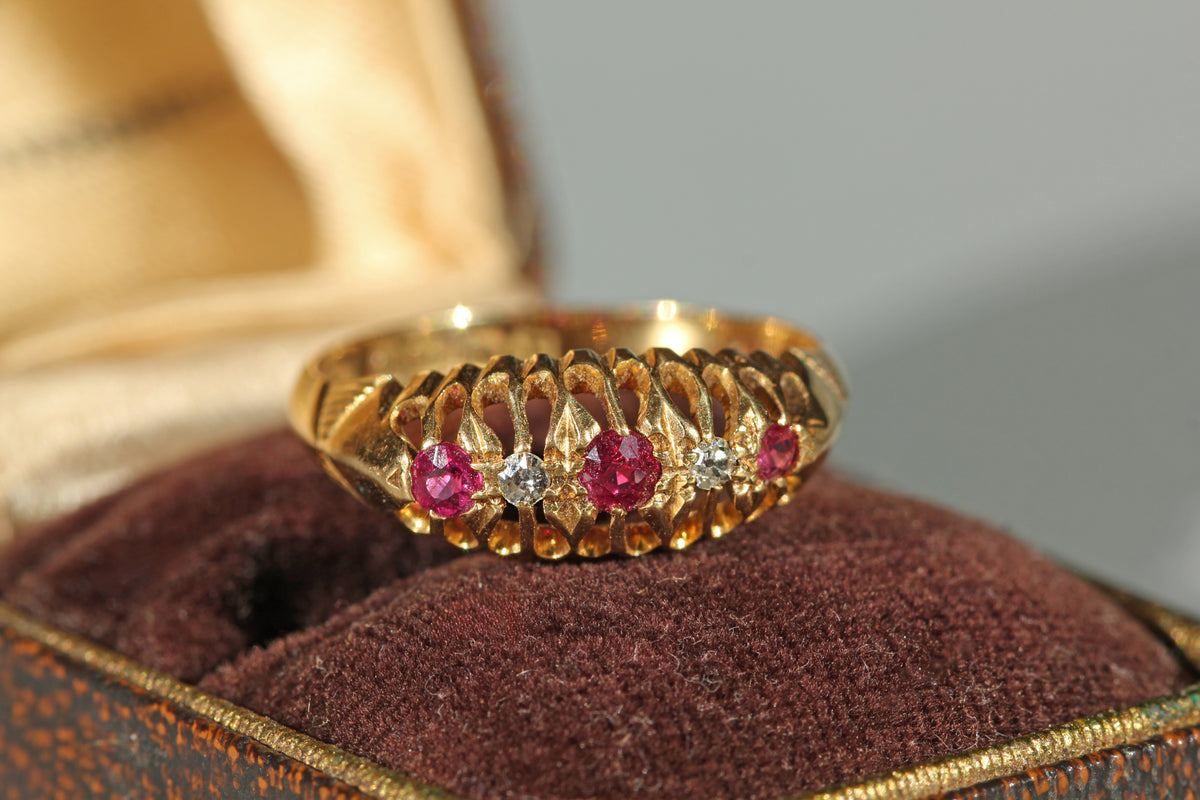 An 18ct yellow gold ring with three pink ruby gemstones and old cut diamonds, displayed on a brown antique velvet ring box.