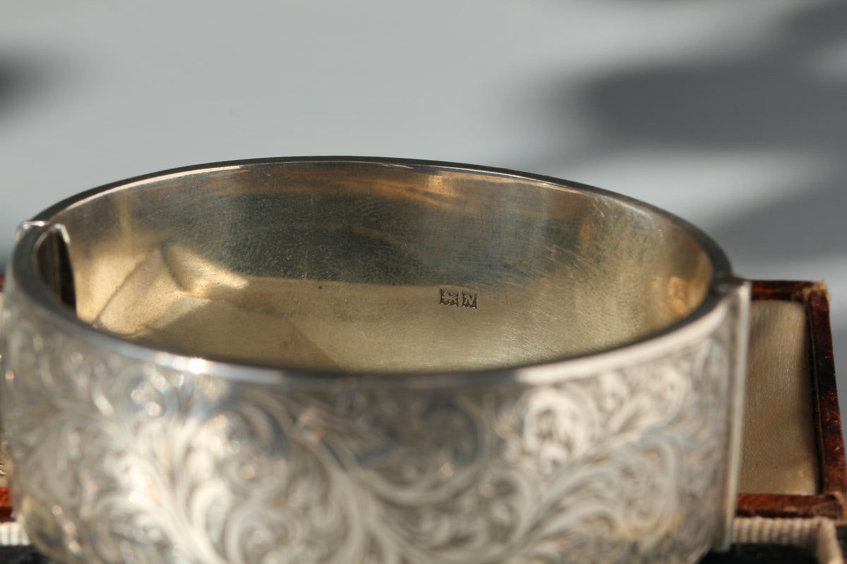 A close-up of a wide silver bangle bracelet with ornate floral engraving on the exterior and a polished silver interior. A small sterling silver hallmark is visible inside the band.