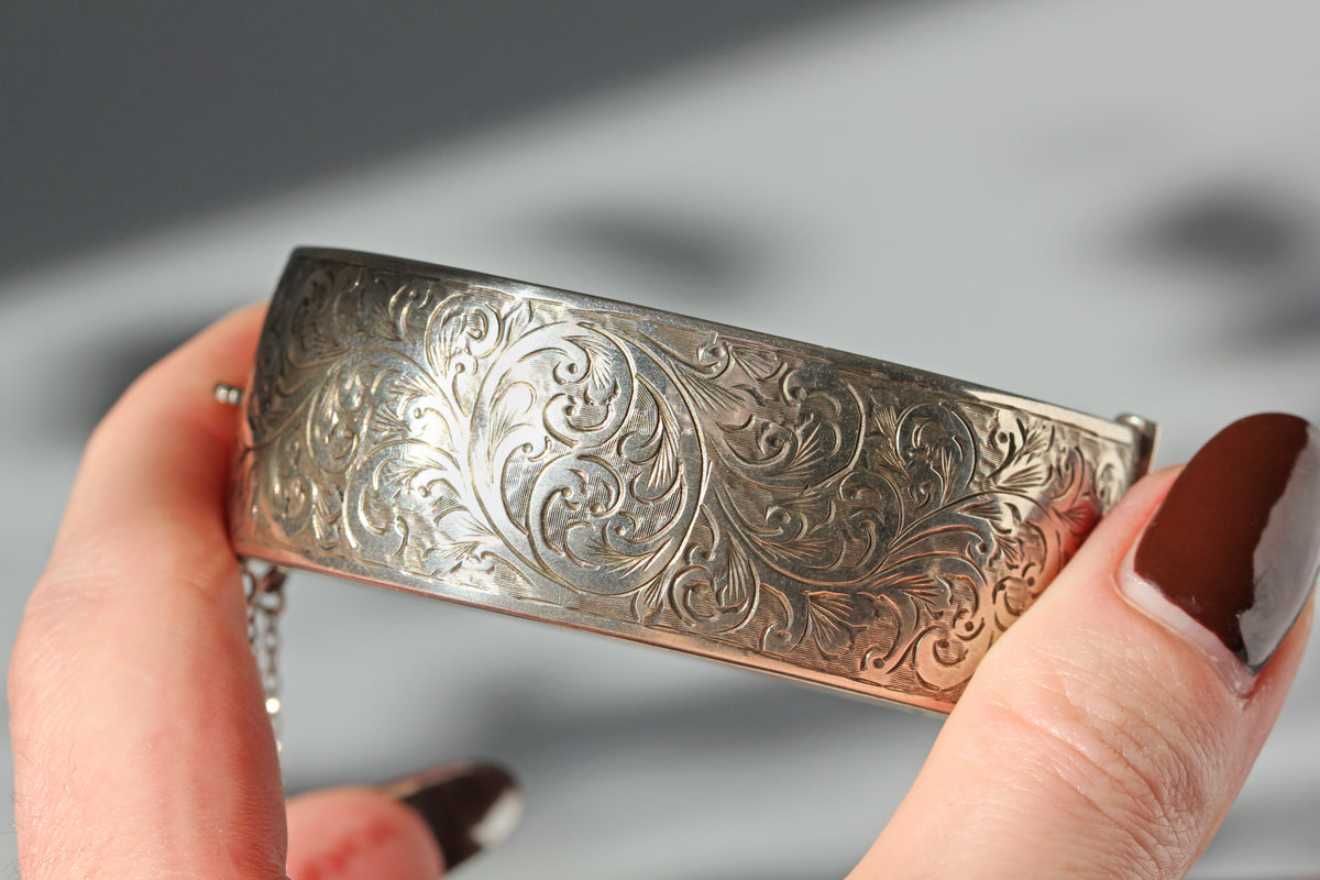 A close-up of a hand holding a wide sterling silver vintage cuff bracelet with intricate floral scrollwork engraving.