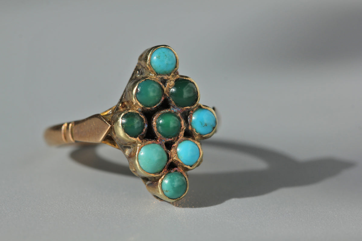 A gold ring featuring a cluster of round turquoise and green gemstones, photographed on a neutral gray background with a soft shadow.