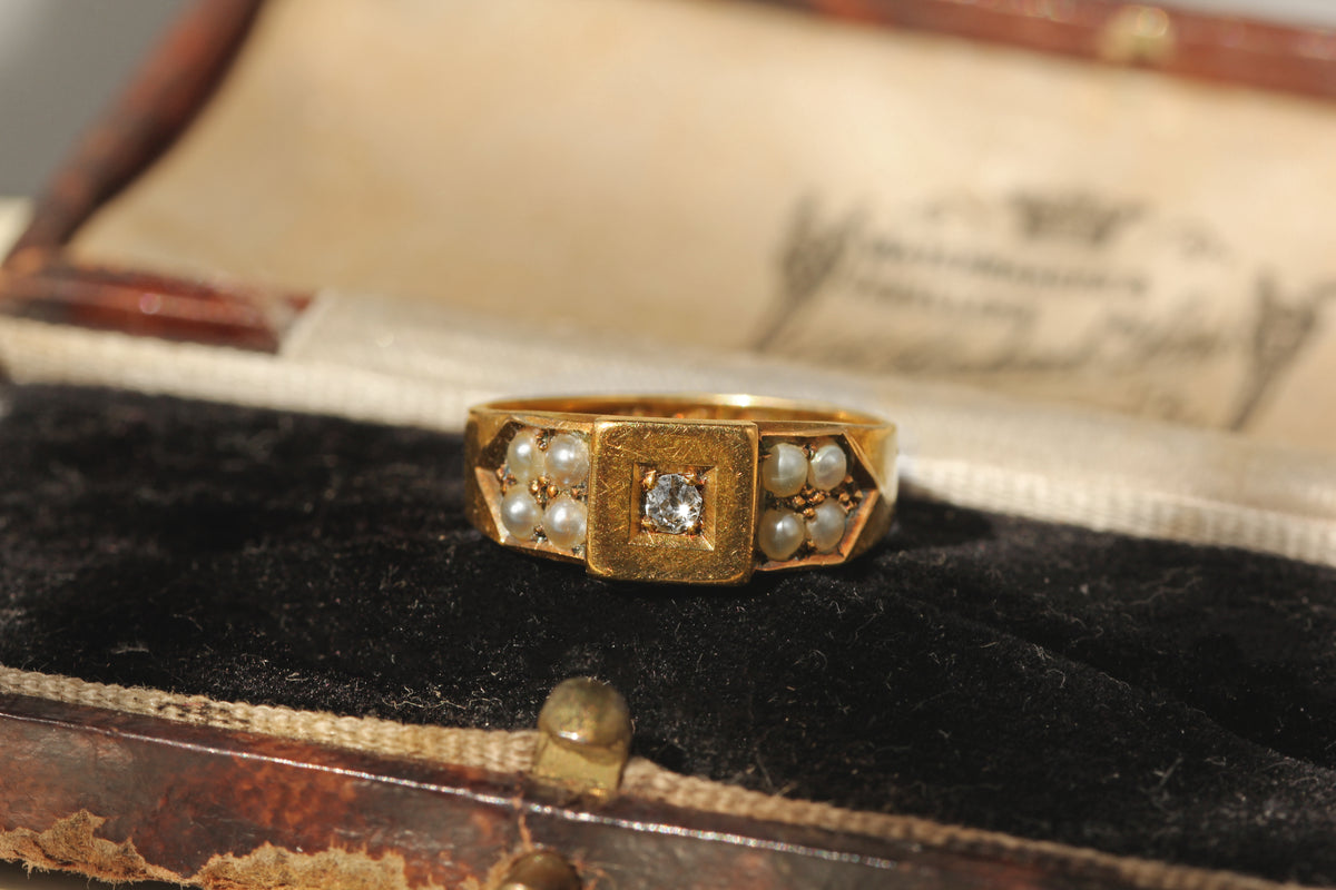 a highly decorative victorian antique band ring, with a geometric face, an old mine cut diamond solitaire and eight seed pearls displayed on an antique jewellery box