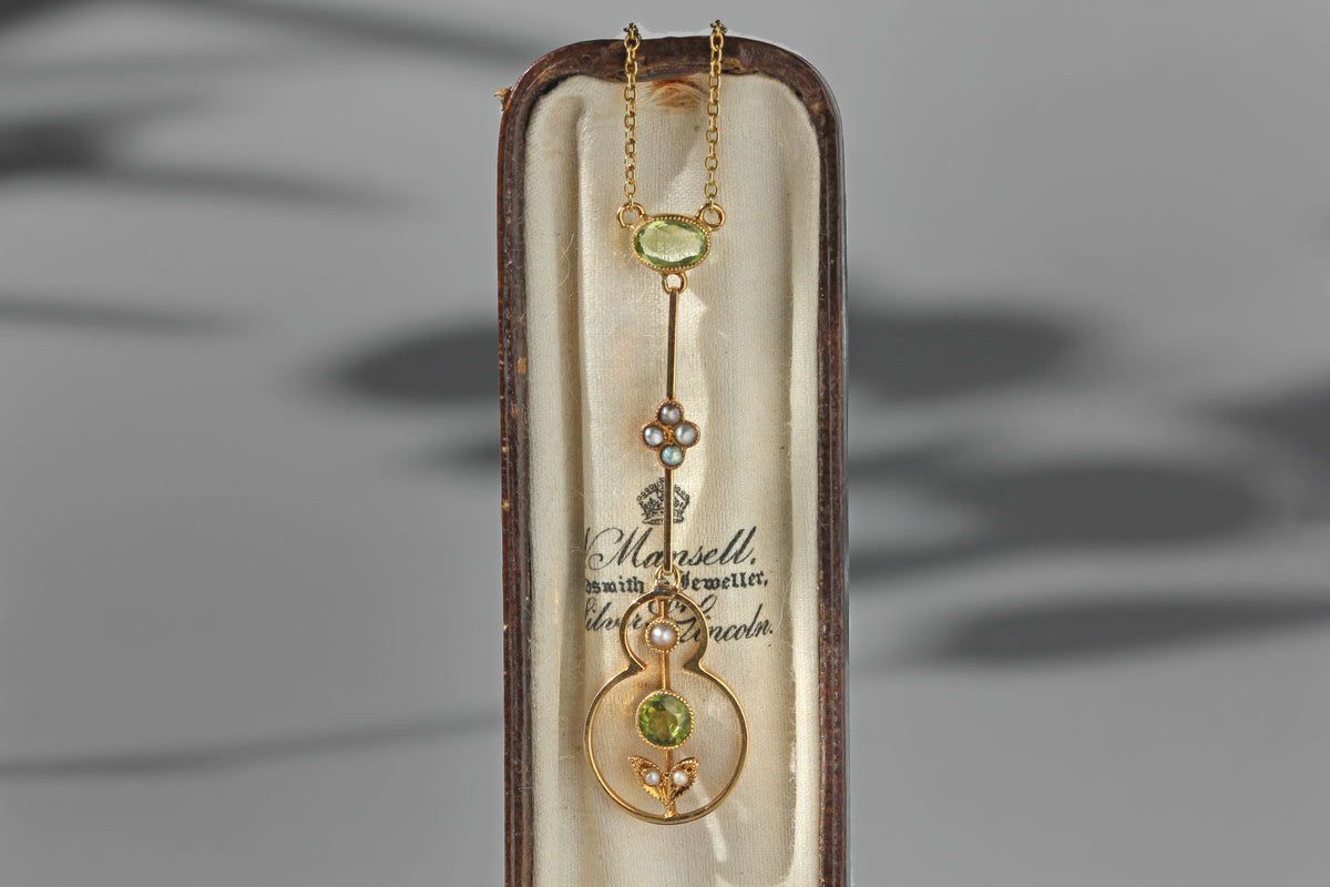 An antique victorian 15ct gold pendant necklace with green peridot gemstone accents, presented inside its original cream colored display box. The pendant features a gold chain, a floral motif with small seed pearls, and a circular gold frame with a green peridot gemstone at the center.