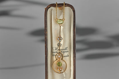 An antique victorian 15ct gold pendant necklace with green peridot gemstone accents, presented inside its original cream colored display box. The pendant features a gold chain, a floral motif with small seed pearls, and a circular gold frame with a green peridot gemstone at the center.