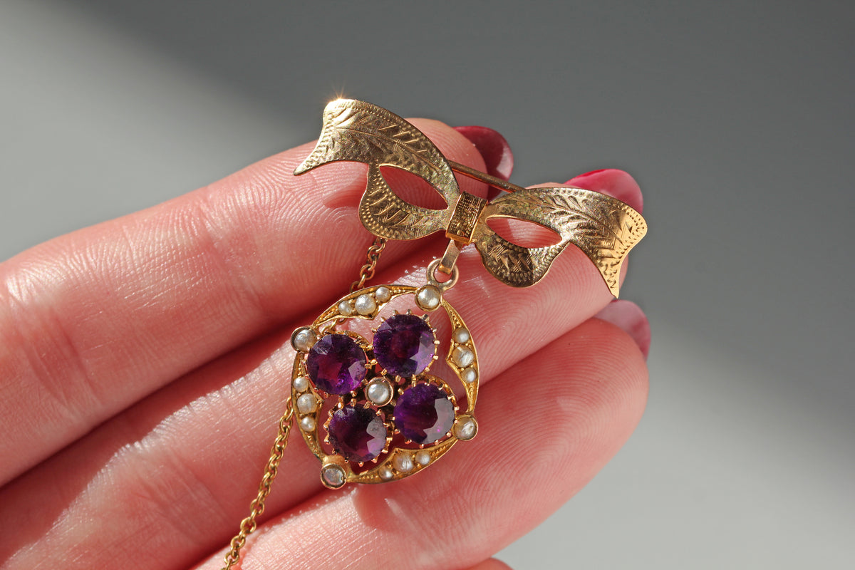 Vintage 9ct gold bow brooch with suspended antique amethyst and pearl floral pendant which dates to the victorian era, held by a persons hand