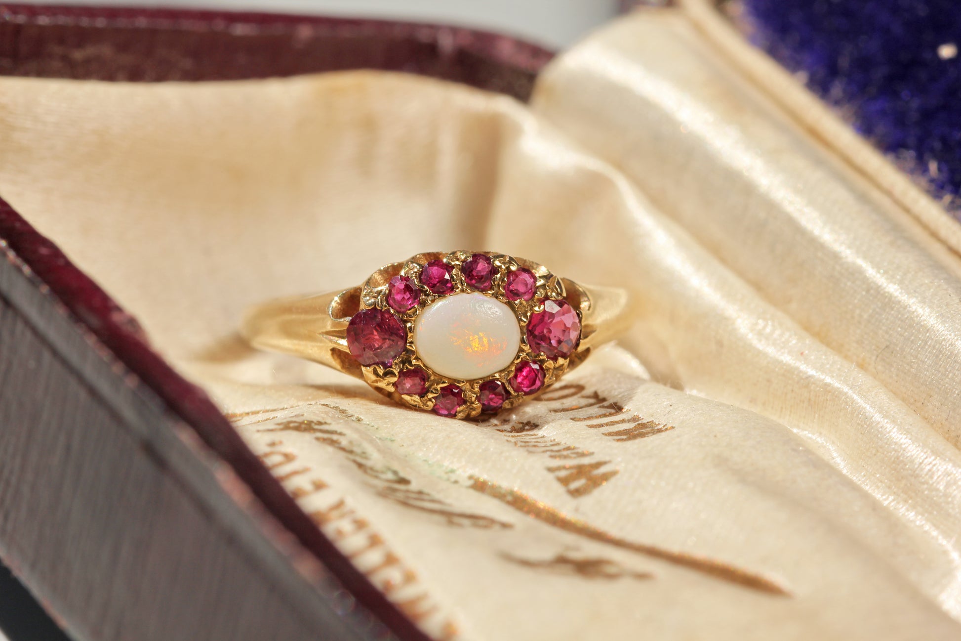 A vintage gold ring with a central white opal surrounded by ruby gemstones, displayed inside an antique velvet jewelry box