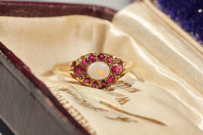 A vintage gold ring with a central white opal surrounded by ruby gemstones, displayed inside an antique velvet jewelry box