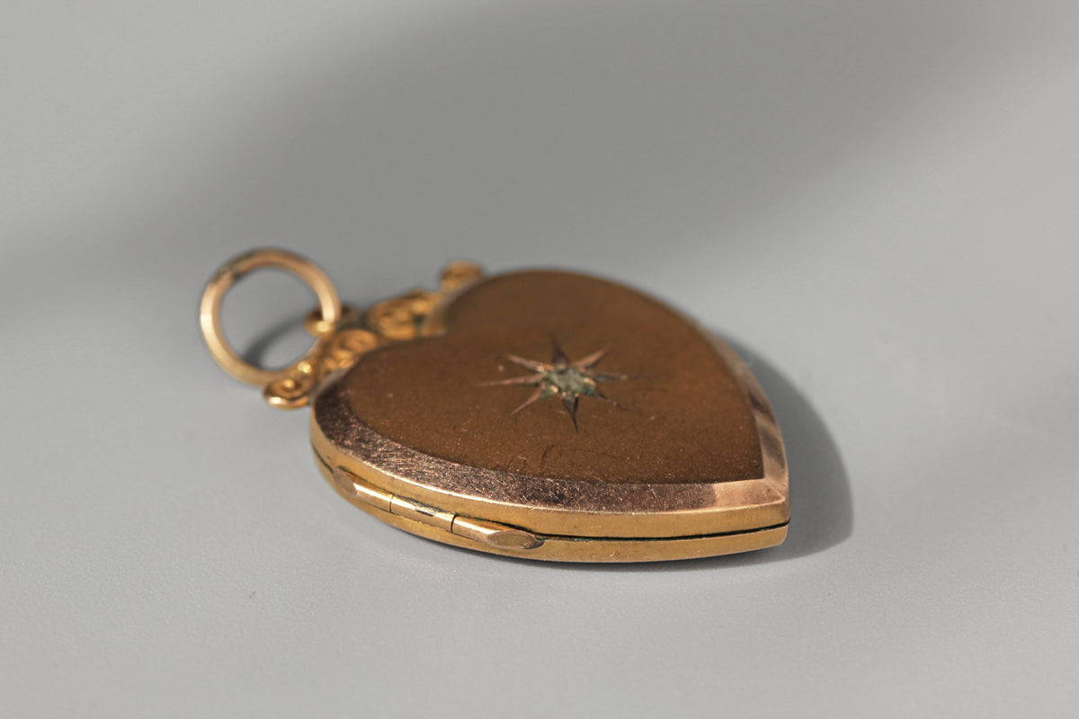 A vintage gold heart-shaped locket pendant with a brown inlay, featuring a central starburst motif and a small loop at the top, shown on a neutral gray background.