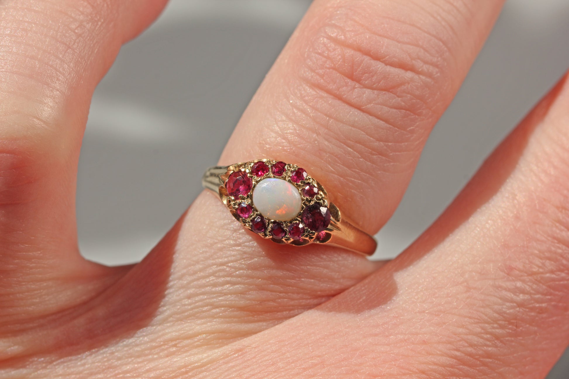 A close-up of a hand wearing a vintage-style ring with a white opal center stone surrounded by a halo of red gemstones on a gold band.